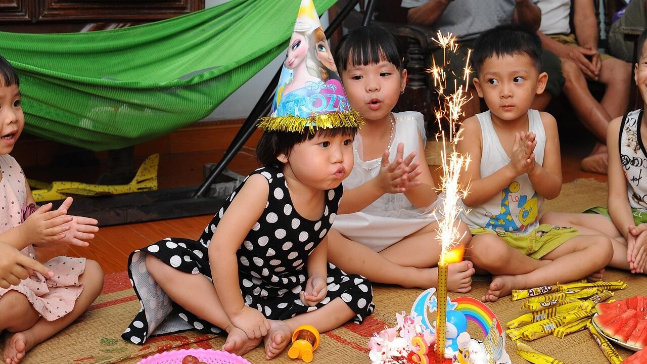 Mädchen mit Partyhut pustet beim Kindergeburtstag die Kerze aus, Freunde klatschen