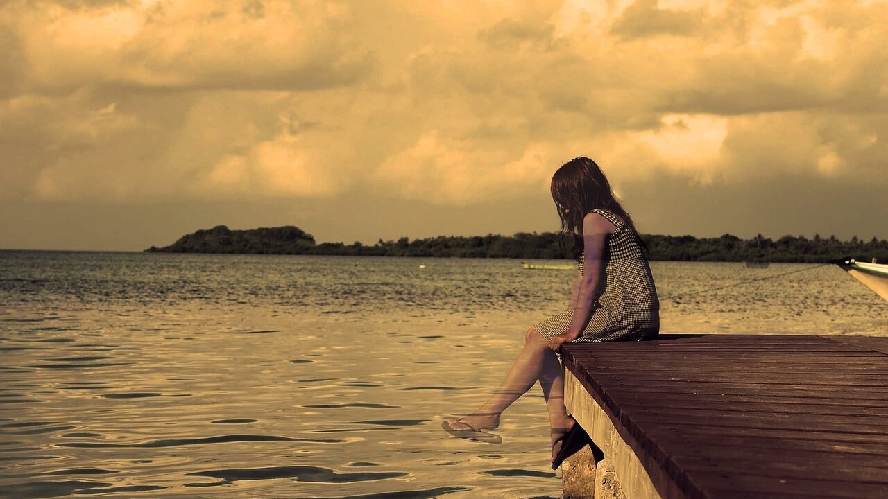 Frau sitzt nachdenklich auf einem Steg am See im Sonnenuntergang